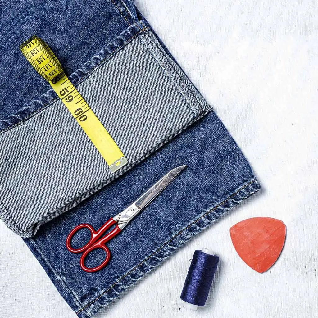 Uma tesoura, uma fita métrica e um par de jeans dispostos sobre uma superfície clara, prontos para um projeto de costura.