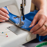 Mulher trabalhando em uma máquina de costura, dedicando-se à confecção de roupas, com materiais variados ao seu redor.